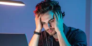 Ung man sitter framför laptop med händerna mot huvudet, ser frustrerad ut – symboliserar osäkerhet kring karriär och framtid.