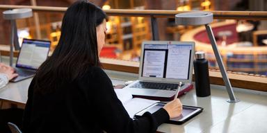 Kvinna studerar med laptop och surfplatta i en biblioteksliknande miljö.