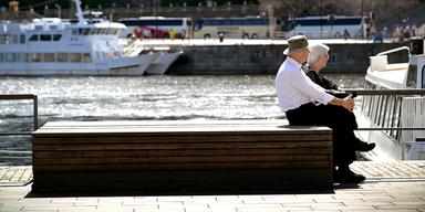 En äldre man och en äldre kvinna som sitter på en bänk vid Grand Hotel i Stockholm. I bakgrunden syns skärgårdsbåtar